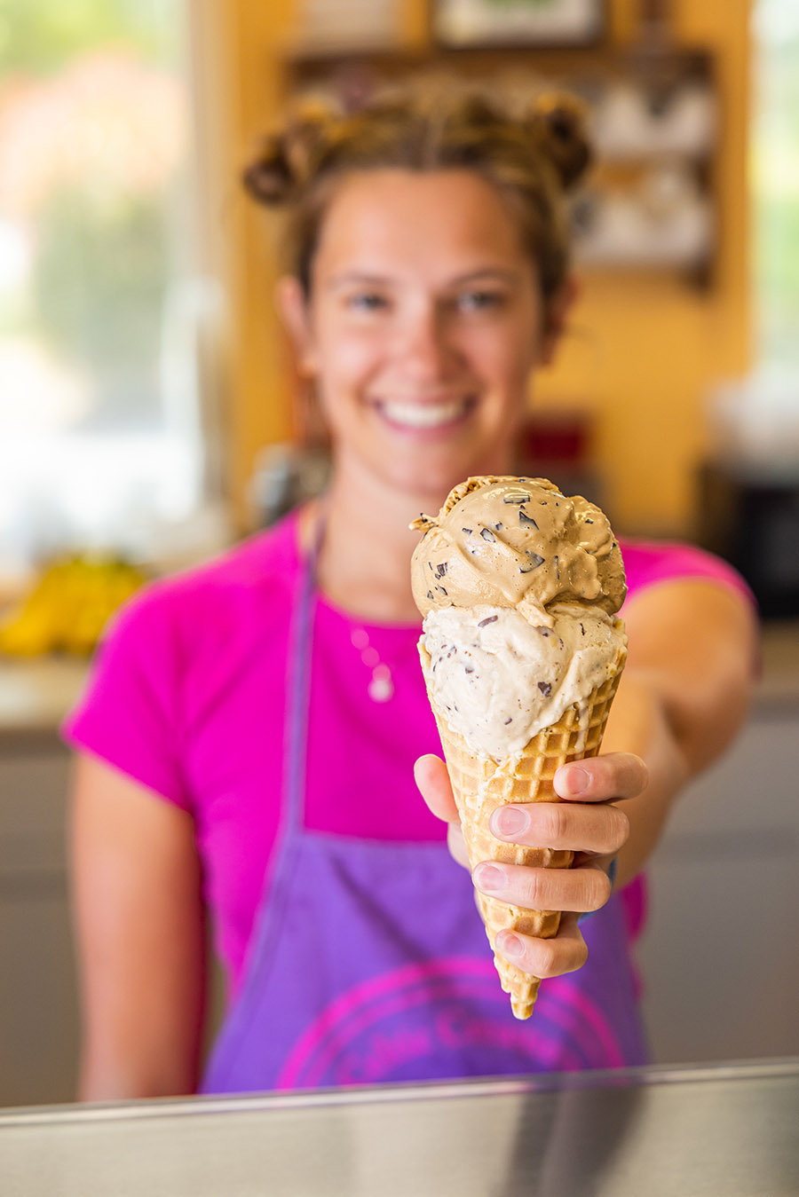 teenage girl wearing an apron holding out ice cream cone with 2 scoops toward the camea